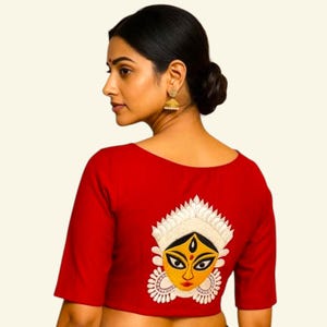 May include: A red blouse with short sleeves and a scoop neckline. The back of the blouse features an embroidered design of a deity's face with a yellow face, black eyes, and white and gold accents. The blouse is made of a smooth fabric.