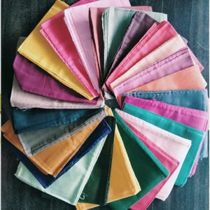 May include: A circular arrangement of folded handkerchiefs in various colours. The palette includes pinks, yellows, greens, blues, and purples. The image is shot from above, highlighting the spectrum of shades. The handkerchiefs are neatly folded.