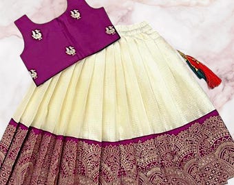 Könnte beinhalten: Ein zweiteiliges Outfit mit einem ärmellosen, tiefvioletten Oberteil mit gestickten Details und einem cremefarbenen Rock. Der Rock hat einen dekorativen Saum mit einem bordeauxroten und goldenen Muster. Ein Quastendetail hängt am Rock.