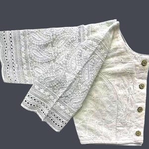 Könnte beinhalten: Weiß bestickte Bluse mit Paisley- und Blumenmustern. Die Bluse hat lange Ärmel mit Lochmuster und eine Knopfleiste vorne. Der Stoff scheint leicht und für warmes Wetter geeignet zu sein.