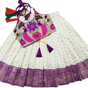 Peut inclure: Ensemble indien traditionnel. La jupe crème est ornée de pois dorés et d'une bordure violette avec des motifs dorés. Le haut beige présente des motifs floraux et paons roses et violets. Comprend un couvre-chef assorti.
