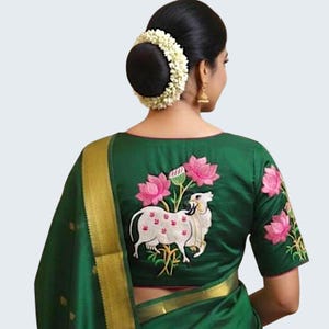 Könnte beinhalten: Eine grüne Bluse mit einem gestickten Design mit einer weißen Kuh mit rosa Akzenten und Lotusblüten. Die Bluse ist Teil eines traditionellen indischen Saris, mit einem sichtbaren Goldrand. Das Haar der Frau ist zu einem Dutt frisiert und mit weißen Blumen geschmückt.