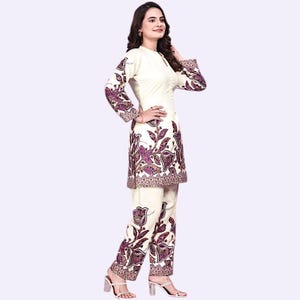 May include: Cream-colored two-piece outfit featuring a tunic and pants. The tunic has long sleeves and a mandarin collar. Both pieces are adorned with a floral pattern in shades of purple and brown. The outfit is worn with silver heeled sandals.