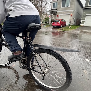 May include: A person riding a black bicycle on a wet street. The bicycle has a black seat, tires, and fenders. A red car is parked in front of a two-story house with a red roof. The sky is overcast.