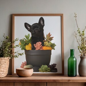 Op de afbeelding: Een ingelijste print met een zwarte Chihuahua-puppy in een donkergrijze bloempot gevuld met kleurrijke vetplanten en bloemen. Het kunstwerk heeft een licht houten frame en wordt tentoongesteld op een houten oppervlak.