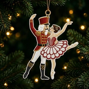 Op de afbeelding: Een hangend ornament met een geborduurde notenkraker-soldaat in een rood uniform en witte broek die danst met een ballerina in een rood-witte tutu. Het ontwerp bevat gouden accenten, zwarte laarzen en een gouden lus aan de bovenkant om op te hangen.