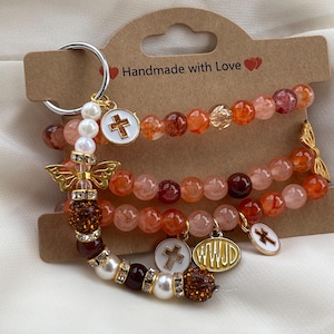 May include: A set of three beaded bracelets in shades of orange, red, and white, featuring a silver keyring attachment. The bracelets include white pearl-like beads, clear crystals, and charms shaped like crosses and an oval with the text WWJD.