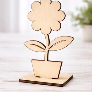 Pode incluir: Uma escultura de flores de madeira. A flor, as folhas e o vaso são cortados em madeira clara. A flor assenta sobre uma base retangular, criando um efeito 3D. O fundo está desfocado.