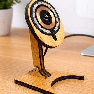May include: Wooden wireless charger stand with a circular charging pad. The pad has a copper centre and a black and white outer ring. The stand is made of light-coloured wood and has a curved base. The stand is designed to hold a phone.