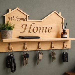 Puede incluir: Llavero de madera con forma de casa con las palabras "Welcome Home" talladas. Tiene un estante, ganchos para llaves, una pequeña planta y un objeto decorativo. Un teléfono móvil descansa sobre el estante.