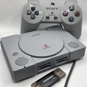May include: A gray PlayStation console with a matching controller and a black USB drive. The console has the PlayStation logo and the word "SONY" printed on it. The controller has directional buttons and action buttons.