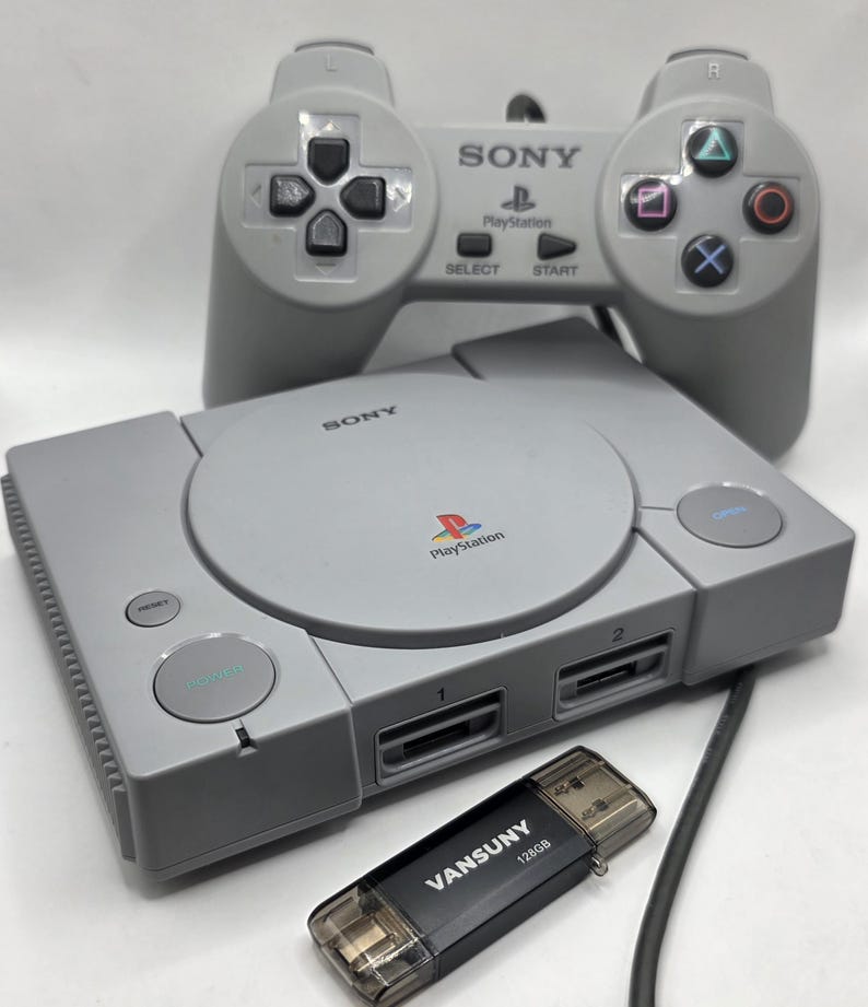 May include: A gray PlayStation console with a matching controller and a black USB drive. The console has the PlayStation logo and the word "SONY" printed on it. The controller has directional buttons and action buttons.