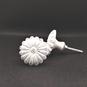 May include: Two white daisy-shaped cabinet knobs with a distressed finish. The knobs have a detailed floral design and a metal screw for installation. The knobs are likely used for furniture or decorative purposes.
