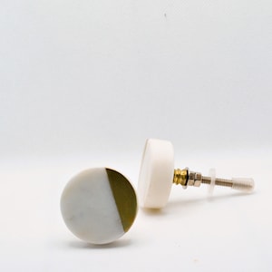 May include: Two decorative cabinet knobs. One knob is white and gold, the other is white. The knobs have a round shape with a metal screw and hardware. The knobs are on a white background.