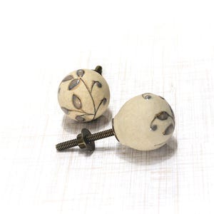May include: Two off-white ceramic cabinet knobs with a leaf design in gray and brown. The knobs have a round shape and are attached to a metal screw. The knobs are designed for furniture and home decor.