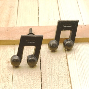 May include: Two cast iron cabinet knobs shaped like musical eighth notes. The knobs are dark brown with a slightly textured surface and a round base. They are set against a light wood background.
