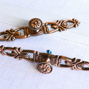 May include: Two ornate bronze-colored cabinet pulls with floral and scrollwork details. Each pull has a central knob with a flower design and a decorative backplate. One pull has a blue knob.