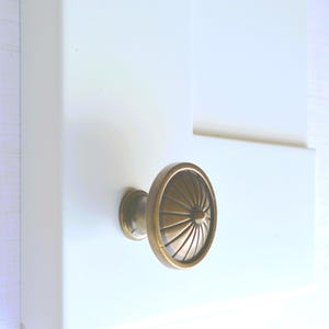 May include: A bronze-colored cabinet knob with a circular, ridged design. The knob is mounted on a white cabinet door, showcasing a classic hardware style. The knob's design features a series of radiating lines, adding a touch of elegance to the cabinet.