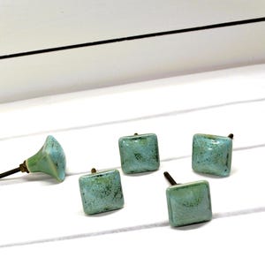May include: A collection of five square ceramic cabinet knobs in shades of turquoise and green. One knob is a cone shape. The knobs have a distressed, textured finish and brass hardware. The knobs are arranged on a white surface.