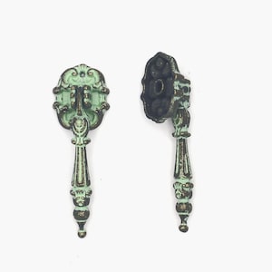 May include: Two antique-style drawer pulls with a distressed finish. The pulls are a pale green with bronze accents, featuring ornate detailing on the handle and backplate. The handles are vertically oriented, with a decorative design.