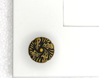 May include: A round, antique brass-colored cabinet knob featuring a detailed, embossed leaf pattern radiating from a central point. The knob is mounted on a flat, white surface with a visible corner edge, showcasing its metallic finish and texture.