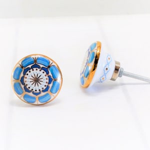 May include: Two decorative ceramic cabinet knobs. The knobs feature a white base with a blue and gold floral design. The knobs have a gold rim and a silver screw. The knobs are on a white surface.