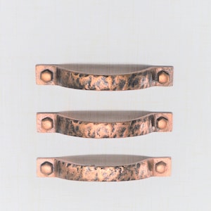 May include: Three copper-colored cabinet pulls with a hammered texture. Each pull has a curved handle and is secured with hexagonal bolts at each end. The pulls are arranged vertically against a white background.