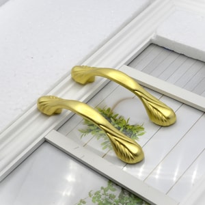 May include: Two gold-colored cabinet handles with a leaf-like design. The handles are curved and have a smooth, metallic finish. They are set against a white background, suggesting a home decor or hardware theme.