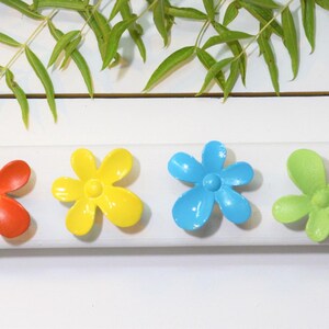 May include: Four small flower-shaped hair clips in bright colors, including red, yellow, blue, and light green. Each clip features five rounded petals and a central circular detail, arranged in a horizontal row on a white surface.