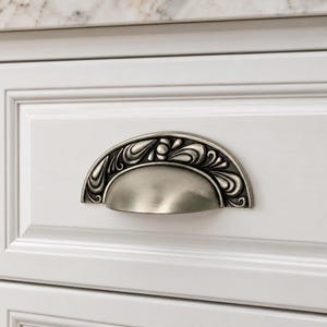 May include: A semi-circular cup drawer pull with an antique silver finish. The hardware features a smooth, rounded handle section at the bottom and an ornate, raised decorative border along the top edge with swirling, leaf-like patterns.