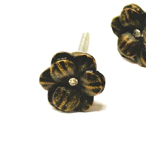 May include: Close-up of two bronze-colored flower-shaped cabinet knobs with a small, clear crystal in the center of each. The knobs have a distressed finish and a threaded post for installation. The background is white.