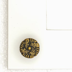 May include: A decorative cabinet knob with a floral design. The knob is made of a dark brass-colored metal and is mounted on a white surface. The knob has a circular shape with a detailed, swirling pattern.