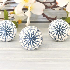 May include: Three white ceramic cabinet knobs with a navy blue compass rose design. The knobs are round and have a glossy finish. Decorative floral accents are in the background.