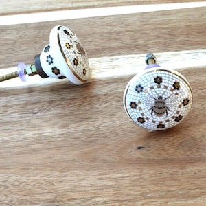May include: Two decorative ceramic cabinet knobs. The knobs are white with a mosaic pattern, gold trim, and a bee design. The hardware includes a bolt, nut, and washer. The knobs are on a wooden surface.