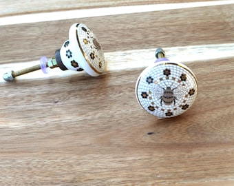 May include: Two decorative ceramic cabinet knobs. The knobs are white with a mosaic pattern, gold trim, and a bee design. The hardware includes a bolt, nut, and washer. The knobs are on a wooden surface.