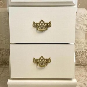 May include: A white two-drawer cabinet with ornate gold-colored drawer pulls. The cabinet has a simple design with a slightly curved base. The drawers are rectangular and the cabinet appears to be made of wood.