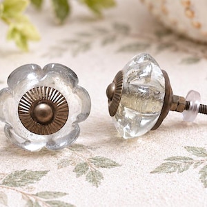 May include: Two flower-shaped cabinet knobs made of clear glass with a faceted surface. Each knob features a central circular bronze-toned metal plate with radial grooves and a threaded metal bolt extending from the back for installation.