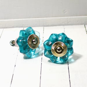 May include: Two turquoise glass flower-shaped cabinet knobs with gold-colored metal accents. The knobs are set against a white wooden background, showcasing their decorative design. These knobs are ideal for adding a touch of elegance to furniture.