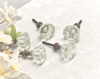 May include: Five clear glass cabinet knobs with faceted, flower-like designs. Each knob has a dark metal screw and a brass-colored base. The knobs are arranged on a white surface, with white flowers in the background.