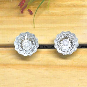 May include: Two clear, faceted glass knobs with a flower-like design. The knobs are round and have a detailed, sparkling appearance. They are set against a light wood background, suggesting a vintage or decorative theme.