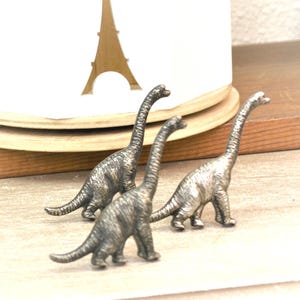 May include: Three small, silver-colored dinosaur figurines with long necks and detailed textures. The dinosaurs are positioned on a light-colored surface, with a white background and a wooden shelf visible. The figurines are a decorative item.