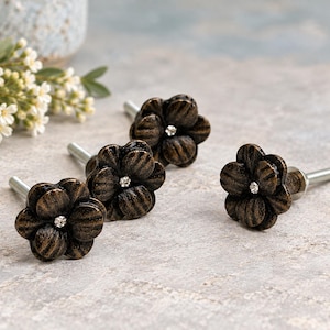 May include: Four flower-shaped cabinet knobs with a dark bronze-toned finish and a textured surface. Each knob features a small, clear, circular crystal embedded in the center and a silver-toned metal bolt extending from the back.
