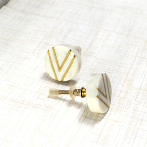 May include: Two white cabinet knobs with gold chevron designs. The knobs have a round shape and are made of a smooth material. The hardware includes a screw and a brass-colored base. These decorative knobs are suitable for furniture.