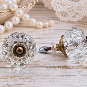 May include: Two round cabinet knobs featuring clear, faceted glass tops with a textured, diamond-like pattern. Each knob has a circular antique brass-toned metal base, a threaded metal bolt, a small plastic washer, and a metal nut.