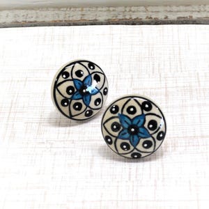 May include: Two ceramic cabinet knobs with a floral design. The knobs are round, with a cream-colored base, black outlines, and a blue flower in the center. The knobs are approximately 1.5 inches in diameter.