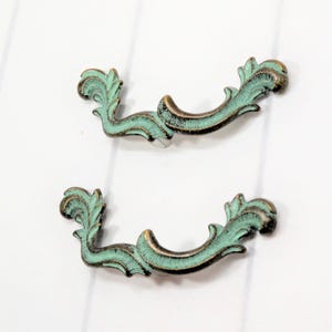 May include: Two antique-style drawer pulls with a distressed teal finish and ornate, leaf-like detailing. The handles have a curved design and are made of metal. These decorative pulls are ideal for furniture restoration projects.
