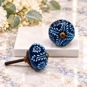 May include: Two round ceramic cabinet knobs featuring a deep navy blue base with light blue floral patterns and a central brass-toned metal accent. Each knob includes a threaded metal bolt extending from the back for installation purposes.