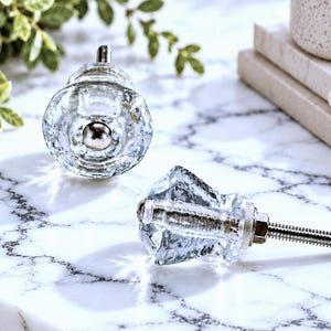 May include: Two clear glass cabinet knobs with a faceted, diamond-like shape and a silver-toned metal center. One knob is shown from the front, while the other is positioned on its side, revealing a long, threaded metal bolt and a nut.