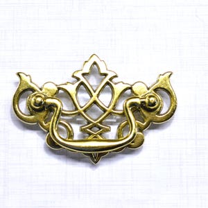 May include: Ornate brass drawer pull with a curved handle and decorative filigree design. The hardware features a symmetrical pattern with floral and scroll elements. The pull is a warm gold color and is set against a white background.