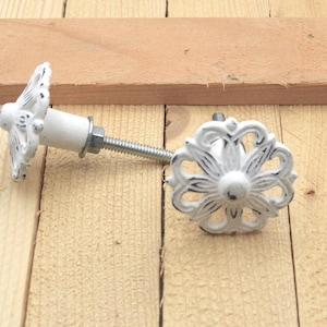 May include: A white, distressed-finish cabinet knob with a floral design. The knob has a long threaded bolt for installation. The hardware is made of metal and is set against a light wood background.